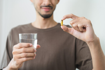Fototapeta premium Dietary supplement, asian young man, male hand holding fish oil pills, drugs medical, take or eat vitamin for treatment skin, hair and nail strengthen with glass of water. Healthcare of person concept