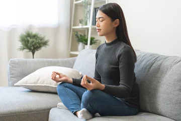 Fotobehang Lotusbloem Beautiful asian young woman, girl hands in calm pose sitting practice meditating in lotus position on sofa at home, meditation, exercise for wellbeing, healthy care. Relaxation, happy leisure people.  © KMPZZZ