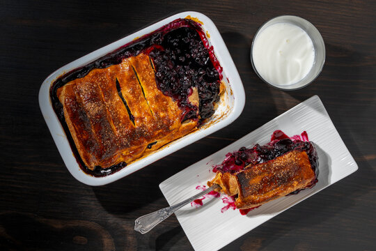 Homemade blueberry cobbler pie with glass of milk, top view, closeup. Delicious baked berry pie in ceramic dish served with slice on plate and glass of fresh milk on dark wooden table