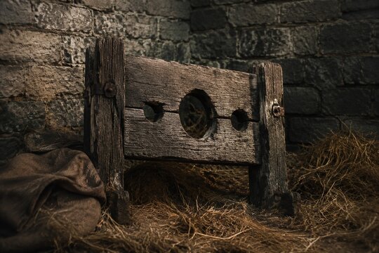 A rough wooden pillory in hay against a stone castle wall.