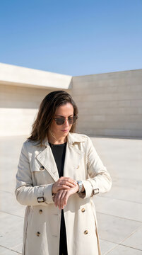 Young stylish woman in a beige trench coat and sunglasses checking time on her watch outdoors