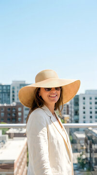 Happy smiling woman wearing a large sun hat and sunglasses standing on a rooftop balcony with city skyline in the background