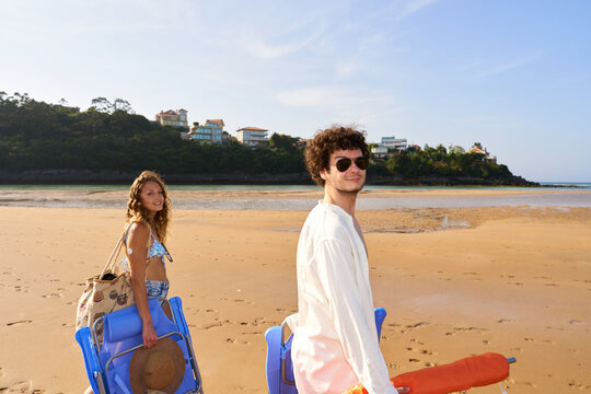 Young couple walking on sandy beach carrying chairs