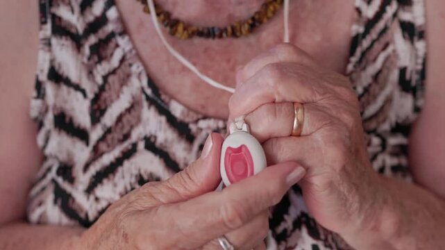 Senior woman with wrinkled hands pressing a red panic button on a medical alert necklace to call for help, ensuring her safety and providing quick access to emergency services from home
