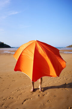Person hiding behind orange beach umbrella on sand