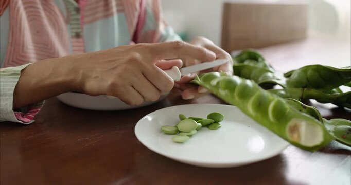 Close up. Asian Woman Hand Peeling Parkia Speciosa , Stink Bean, Bitter Bean, with Knife into White Plate on Table.