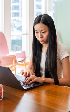 Young Asian Woman Works on Laptop at a Table in a Bright Room