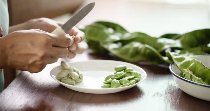Close up. Asian Woman Hand Peeling Parkia Speciosa , Stink Bean, Bitter Bean, with Knife into White Plate on Table.