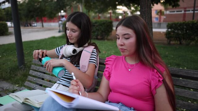 Two focused teenage female friends sitting on a bench in a public park, diligently studying and doing homework together while sharing a drink from a reusable water bottle after school