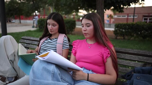Two cheerful adolescent girls collaborating on their homework after school, sitting on a park bench and discussing their notes with enthusiasm while preparing for an upcoming exam
