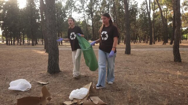 Two dedicated young female volunteers with recycling symbol shirts actively collect plastic and cardboard trash from the ground, putting it into a green garbage bag during a park cleanup event
