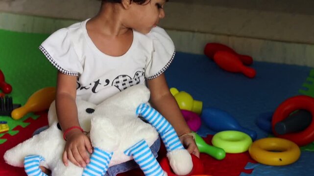 A cute little Indian toddler girl sitting on a colorful puzzle playmat indoors, happily playing with a soft toy, plastic bowling pins, and colorful rings.
