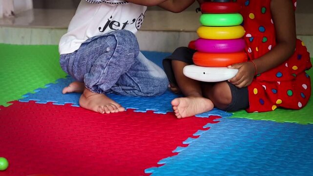 Close-up of two unrecognized Indian children sitting barefoot on a colorful playmat indoors, sharing and playing together with a colorful plastic ring stacker educational toy.

