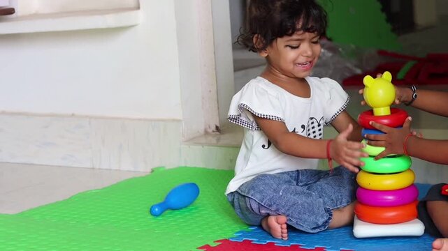 A cute little Indian toddler girl sitting on a colorful playmat indoors, clapping her hands and smiling with joy after successfully completing a colorful plastic ring stacker toy 