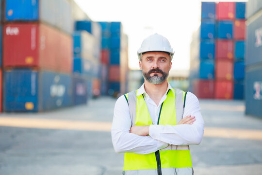 Man manager cross arm. Handsome caucasian male dock foreman worker in white hard hat helmet and high-visibility vest walking in Shipping Cargo Container Terminal Depot