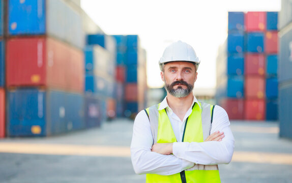 Man manager cross arm. Handsome caucasian male dock foreman worker in white hard hat helmet and high-visibility vest walking in Shipping Cargo Container Terminal Depot