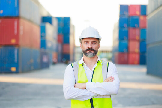 Man manager cross arm. Handsome caucasian male dock foreman worker in white hard hat helmet and high-visibility vest walking in Shipping Cargo Container Terminal Depot