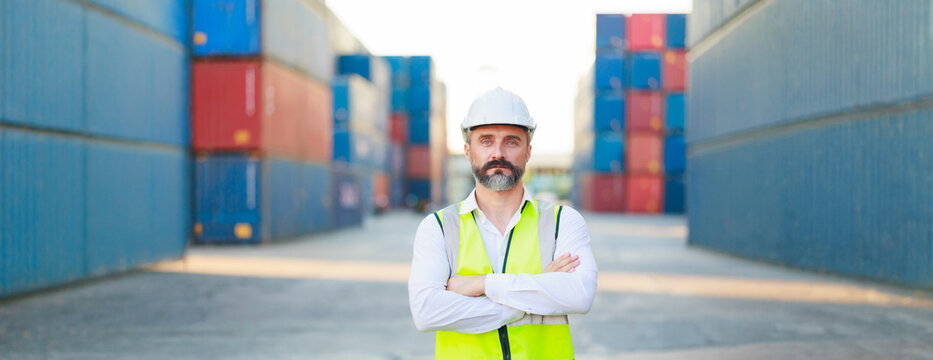 Man manager cross arm. Handsome caucasian male dock foreman worker in white hard hat helmet and high-visibility vest walking in Shipping Cargo Container Terminal Depot