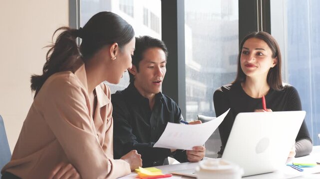 Three business professionals discussing charts and reports while analyzing information on laptop during collaborative office meeting.