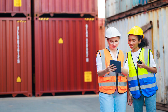 Black female dock worker and caucasian manager control loading containers box on tablet computer from cargo at warehouse container yard. Marine and carrier insurance concept.