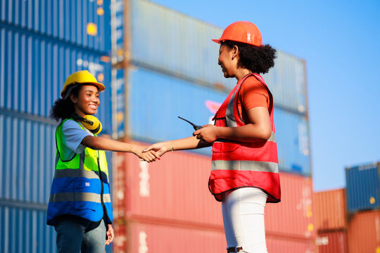 Unity and teamwork concept. Hand shake African woman logistics workers in hardhat helmet and high visibility safety gear at shipping container yard