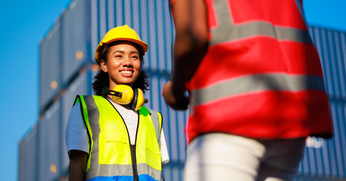 Unity and teamwork concept. Hand shake African woman logistics workers in hardhat helmet and high visibility safety gear at shipping container yard