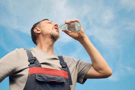 Man in overalls hydrating during work outdoors under hot summer sky
