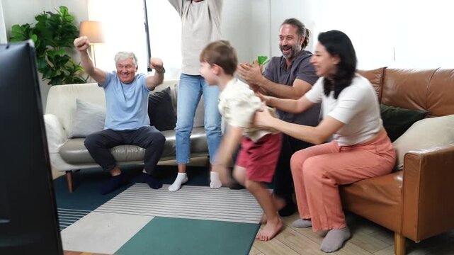 The son, parents, and grandparents sit on the sofa watching TV, cheering on their favorite team, enjoying themselves together in the living room of their home. A family and three-generational concept.