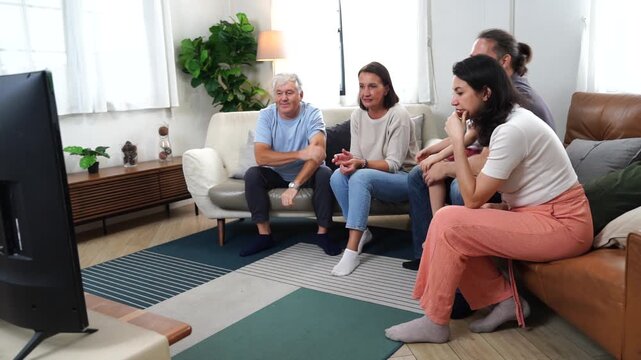 The son, parents, and grandparents sit on the sofa watching TV, cheering on their favorite team, enjoying themselves together in the living room of their home. A family and three-generational concept.