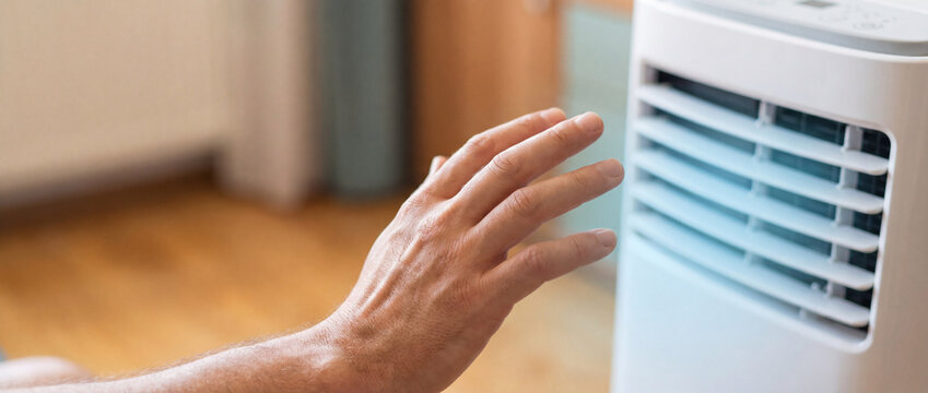 Hand feeling cool air from portable AC unit in a bright modern room, checking the refreshing breeze of a white mobile air conditioner during hot summer days at home for commercial technology