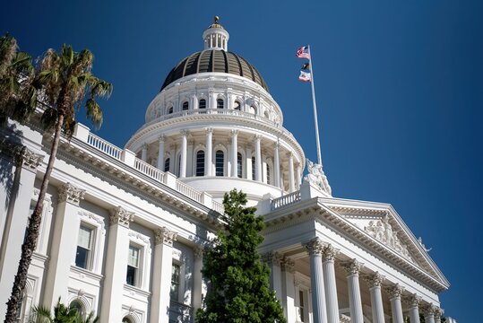 California State Capitol stands tall in Sacramento under a bright blue sky with flags waving and palm trees nearby on a sunny day