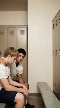 Vertical photo of two teenage boys sitting on bench in locker room. Friend showing solidarity and support. Disappointment and empathy concept