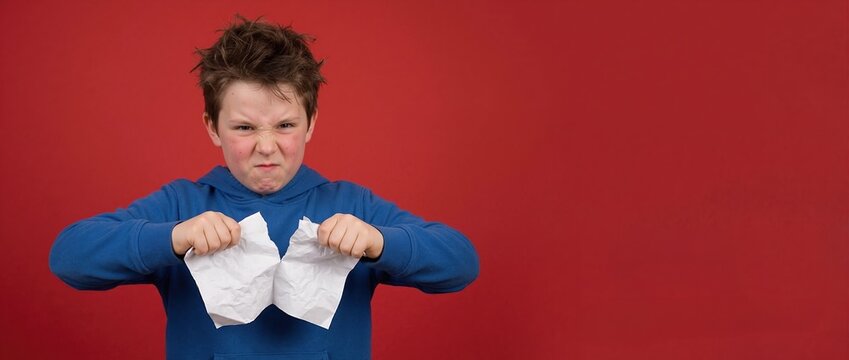Frustrated young boy tearing paper against red background. Angry child in blue hoodie with copy space. Emotional stress and behavior concept. Banner for text