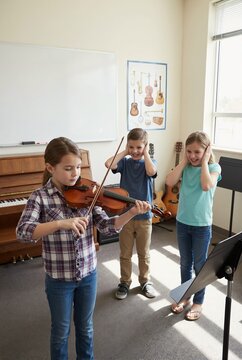 Young girl practicing violin in music classroom. Children in background covering their ears and laughing. Humorous music lesson concept