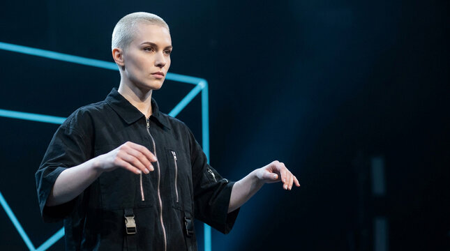 Woman with buzz cut and black jumpsuit gestures with hands on stage