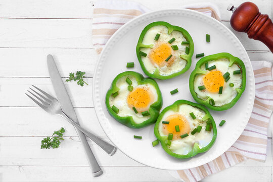 Healthy breakfast concept with eggs in green peppers on a plate. Overhead view table scene on a white wood background.