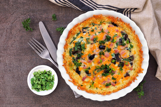 Breakfast quiche with ham, mushrooms, and onions made in a hash brown shell. Overhead view table scene on a dark background.