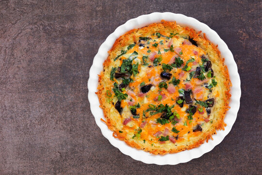 Breakfast quiche with ham, mushrooms, and onions made in a hash brown shell. Top view on a dark background.