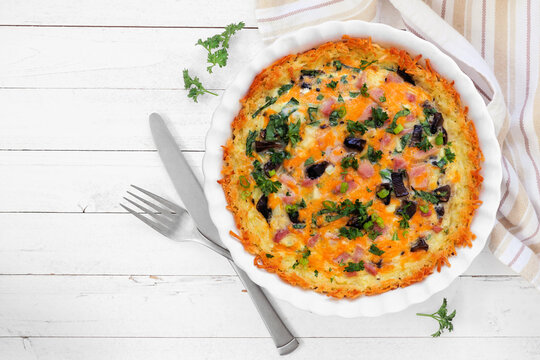 Breakfast quiche with ham, mushrooms, and onions made in a hash brown shell. Above view table scene on a white wood background.