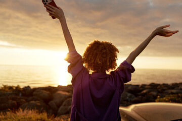 A woman with curly hair stands at sunset by the shore, arms raised in triumph, capturing a carefree, joyful lifestyle moment. The warm glow over water emphasizes freedom and happiness. © SHOTPRIME STUDIO