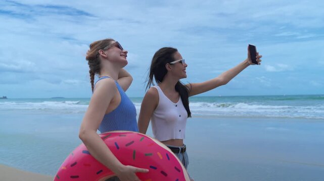 Two young female friends wearing sunglasses taking smartphone selfie and holding colorful inflatable donut pool float at beautiful sandy beach during sunny summer vacation holiday trip.