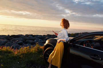 A woman with curly hair stands beside a dark car at sunset by the rocky coastline, feeling contemplative as she gazes at the horizon in a quiet casual lifestyle moment. © SHOTPRIME STUDIO