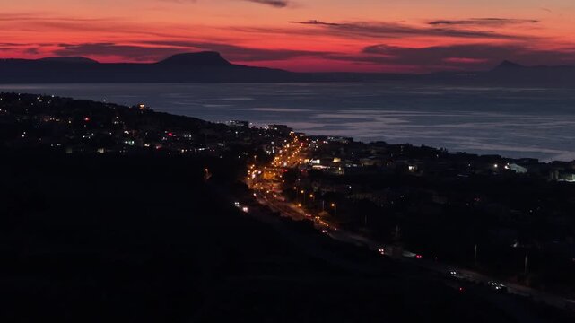 Aerial drone view of a coastal town below a flat-topped mountain across the gulf under a red afterglow, Almyros Gulf, Greece