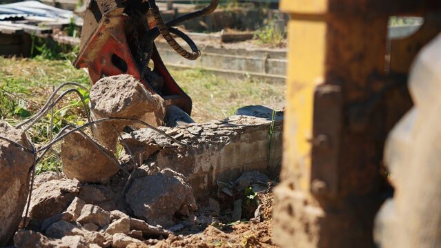 Excavator machine breaking up old concrete foundation or wall with a hydraulic breaker attachment, generating rubble and exposed rebar at a construction site