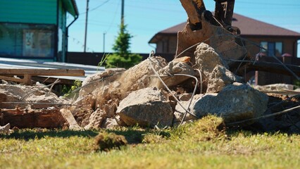 Fototapeta premium Excavator bucket actively breaking reinforced concrete and rebar from a demolished structure, creating rubble on a sunny construction site with a residential background