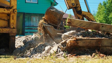 Fototapeta premium Low-angle view of a backhoe bucket holding a reinforced concrete block above demolition rubble, with bent rebar, hanging roots and broken timber at a residential site.