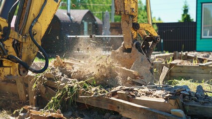 Fototapeta premium Yellow excavator bucket breaking up rubble and wooden debris on a sunny day, clearing the ground for new development and construction work in a suburban area
