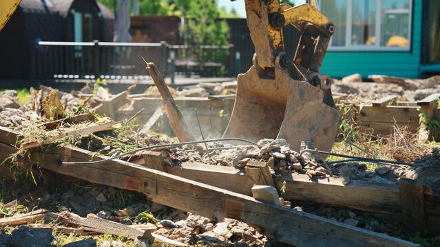 Excavator bucket operating on a construction site, breaking concrete and wood rubble from a destroyed structure, showing active work in progress