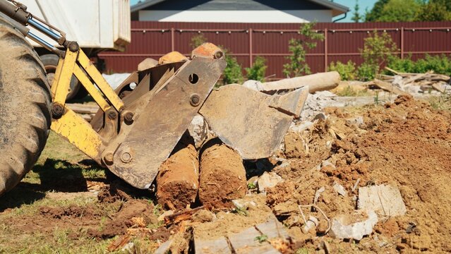 Wide side view of a backhoe loader grapple beside soil, broken concrete and large cylindrical debris during residential demolition site clearance.