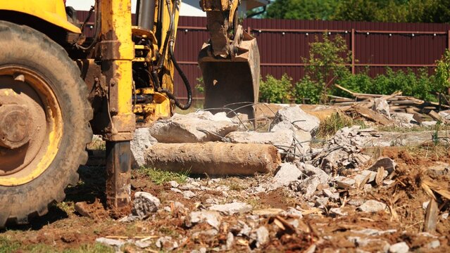 Wide view of a backhoe loader and bucket beside reinforced concrete rubble, exposed steel and broken timber during residential demolition cleanup.